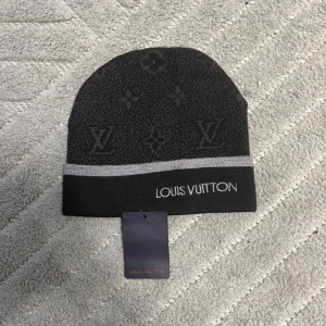 Svart mössa från Louis Vuitton - Svart mössa från Louis Vuitton med ikoniska LV-monogram och diskreta blommor i mörkgrått. Mössan har en grå rand nertill och broderad logga framtill. Tillverkad i mjukt material som passar perfekt för kyliga dagar. Snygg och stilren accessoar för dig som gillar lyx.