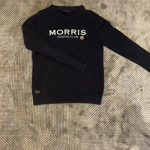 Svart stickad tröja Morris Rowing Club - Snygg svart stickad tröja från Morris Rowing Club med vit broderad logga och liten sköld på bröstet. Tröjan har rund hals, ribbade muddar och är långärmad. Perfekt för dig som gillar klassisk och stilren look.