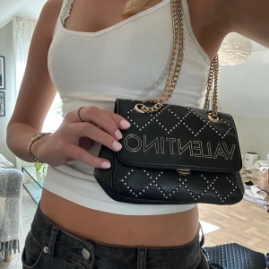 Svart väska från Valentino med gulddetaljer  - PRISET KAN DISKUTERAS!!Snygg svart väska från Valentino som kan bäras på två sätt. Väskor har quiltat mönster och guldfärgade nitar. Väskan har en kedja i guld som axelrem och stor logotyp framtill. Tillverkad i skinn med stilren och modern look, perfekt för dig som gillar statement pieces. Köptes för 2000 kr. Nästan aldrig använd!