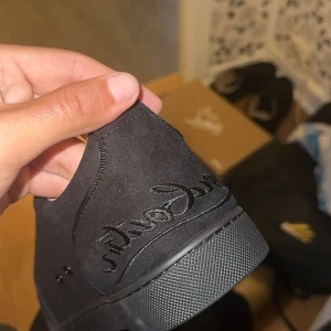 Svarta sneakers från Louboutin - Säljer ett par svarta sneakers från Christian Louboutin med klassisk röd sula och broderad logga på hälen. Skorna har en stilren design i mocka och detaljer i svart, perfekt för dig som vill sticka ut med exklusiva detaljer. Dust bag finns och box inkluderas. Helt oanvända skriv för några frågor eller mer bilder