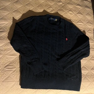 Svart stickad tröja Polo Ralph Lauren - Kabelstickad svart tröja från Polo Ralph Lauren med klassisk rund halsringning och röd broderad logga på bröstet. Tröjan har ribbade muddar och är långärmad, perfekt för kyligare dagar.