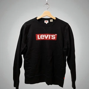Svart sweatshirt från Levi's med logga - Svart sweatshirt från Levi's med klassisk röd och vit logga framtill. Tröjan har rund halsringning och långa ärmar. Tillverkad i mjuk bomull och polyesterblandning, perfekt för en avslappnad och stilren look. 