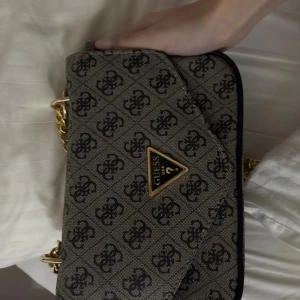 Guess axelväska med monogram och kedja - Snygg axelväska i beige och svart med klassiskt monogrammönster. Väskan har guldfärgad kedja, logga i metall framtill och praktiskt innerfack med dragkedja. Perfekt storlek för det viktigaste och en stilren detalj till din outfit.