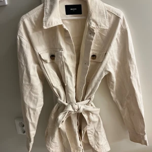 Beige kappa från Bik Bok med bälte - Stilren beige kappa från Bik Bok i bomull med knytskärp i midjan och två bröstfickor med knappar. Jackan har klassisk krage, långa ärmar och snygga detaljer som ger en avslappnad men trendig look.