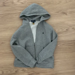 Grå hoodie från Polo Ralph Lauren - Klassisk grå hoodie från Polo Ralph Lauren med dragkedja framtill, två fickor och broderad logga på bröstet. Mjuk insida och huva för extra komfort. Perfekt för chill dagar och enkel att matcha med jeans eller joggers.Säljer denna för att den blivit för liten. Hann knappt att använda den så är inprisip helt ny. Köpte denna för 1195 och säljer den för 449+frakt vilket är ett bra pris enligt mig hör av er vid frågor😊pris kan diskuteras 