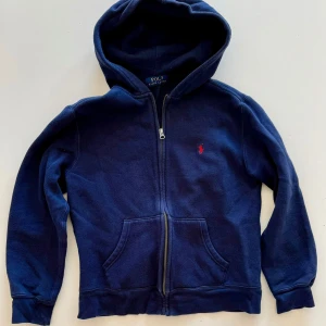 Marinblå hoodie från Polo Ralph Lauren - Marinblå hoodie från Polo Ralph Lauren med dragkedja framtill och klassisk röd logga broderad på bröstet. Tröjan har huva, känguruficka och långa ärmar. Perfekt för en chill och stilren look.