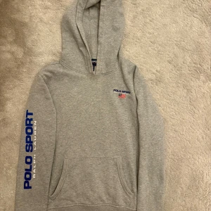 Grå hoodie från Polo Sport Ralph Lauren - Snygg grå hoodie från Polo Sport Ralph Lauren med blå logga på ärmen och liten broderad logga med amerikansk flagga på bröstet. Klassisk känguruficka och huva. Perfekt för en avslappnad och sportig stil.