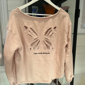 Ljusrosa Odd Molly tröja med fjäril - Superfin ljusrosa tröja från Odd Molly med stort utskuret fjärilsmotiv och texten 'I am with freedom' på ryggen. Tröjan har råa kanter, lång ärm och en avslappnad passform. Perfekt för dig som gillar unika detaljer och mjuka material.