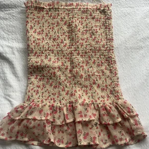 Blommig volangkjol från Bik Bok - Supersöt kort kjol från Bik Bok i ljusbeige med rosa blommor. Kjolen har smock över hela och dubbla volanger längst ner. Perfekt vardags som fest. Använd någon enstaka gång, inga tecken på användning 
