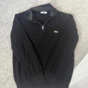 Svart stickad tröja från Lacoste M - Svart stickad tröja från Lacoste i storlek M/ sTröjan har halv dragkedja, ribbad krage med gröna ränder och klassisk broderad krokodil på bröstet. Perfekt för lager-på-lager och chill stil. Den passar både m och s och har blivit mindre i storlek på grund av tvätt