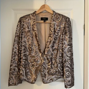 Mango Glittrig kavaj i guld - Säljer en superglittrig kavaj från Mango i guld och silver med paljetter över hela jackan. Kavajen har lång ärm, öppen front och snyggt mönster av snirkliga paljetter. Perfekt för dig som vill sticka ut och älskar statement-plagg!