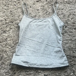 Grått linne med smala band - Säljer ett ljusgrått linne med smala, justerbara axelband. Toppen är figurnära och har en enkel, stilren design som passar perfekt till jeans eller kjol. Mjuk och stretchig bomullsblandning för extra komfort. 