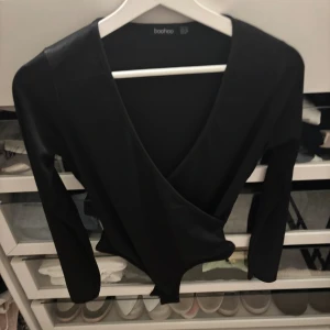 Svart omlottopp från Boohoo - Svart omlottopp från Boohoo med djup v-ringning och långa ärmar. Toppens material är stretchigt och följsamt, vilket ger en snygg passform. Perfekt att styla med jeans eller kjol för en trendig look.