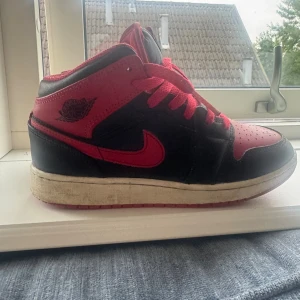 Nike Air Jordan 1 Mid svart/röd 37,5 - Säljer ett par Nike Air Jordan 1 Mid i klassisk svart och röd färg. Skorna har röd yttersula, svarta och röda paneler i skinn, samt röda snören. Ikonisk Jordan-logga på sidan och Nike swoosh. Perfekta för dig som gillar streetwear och sneakers med attityd.