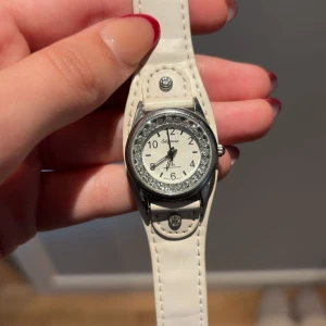 Vit klocka med strass från Cidarenia - Snygg klocka från Cidarenia med vit läderrem och rund urtavla. Runt boetten sitter glittriga strasstenar som ger extra bling. Urtavlan är vit med svarta siffror och detaljer. Remmen har dekorativa nitar med strass för en cool touch.