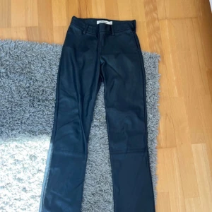 Svarta skinnbyxor Carin Wester 34 - Svarta skinnbyxor från Carin Wester i storlek 34. Byxorna har raka ben, bälteshällor och sidodragkedja. Snygg minimalistisk look med fickdetaljer bak. Perfekta för dig som gillar stilrena och trendiga plagg.