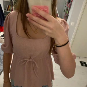 Ljusrosa blus med knyt i midjan - Supersöt ljusrosa blus med puffärmar och fyrkantig ringning från lager 157. Blusen har knyt i midjan och är tillverkad i ett lätt, strukturerat tyg som ger en luftig känsla. Perfekt för vår och sommar när du vill ha något både gulligt och bekvämt. Använd enstaka gång. 