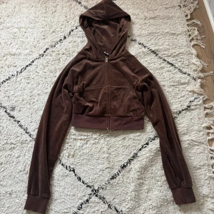 Brun croppad hoodie i velour - Mysig brun croppad hoodie i mjuk velour med huva och dragkedja framtill. Långa ärmar och bred mudd vid midjan ger en snygg passform. Perfekt för dig som gillar en chill och trendig stil.