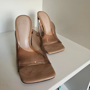 Transparenta klackar från Zara - Säljer ett par trendiga transparenta klackar från Zara i beige/brun ton. Skorna har fyrkantig tå, bred genomskinlig rem och hög blockklack i plast. Perfekta för dig som vill sticka ut med en modern och edgy look.