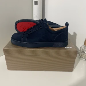 Mörkblå mockasneakers från Louboutin - Snygga mörkblå sneakers i mocka från Christian Louboutin med klassisk röd sula. Skorna har rund tå, platt sula och snörning. Perfekta för dig som vill ha en stilren och lyxig look med en twist.