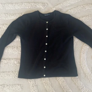 Svart kofta med knappar - En klassisk svart kofta från Daniels i storlek M. Koftan har rund halsringning, långa ärmar och vita knappar längs hela framsidan.