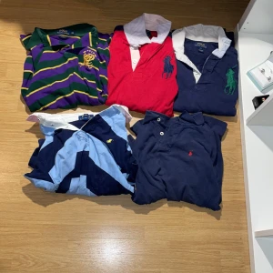 Polo Ralph Laurent pikér bulk x5 - Flera klassiska pikétröjor från Polo Ralph Lauren. En röd med vit krage och stor blå logga, en randig i grönt, lila och gult, samt flera marinblå med olika färger på loggan. Alla har krage och knappar framtill, tillverkade i bomull. Perfekta för en preppy look.