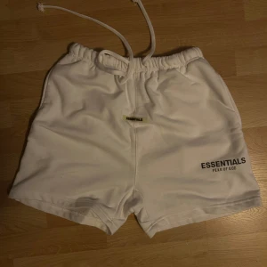 Vita Essentials Fear of God shorts - Säljer ett par vita shorts från Essentials Fear of God med svart logotyptryck på benet och en liten Essentials-tag framtill. Modellen har elastisk midja med snörning och en bakficka. Perfekta för chill eller streetwear. Materialet är mjuk bomull. Jag skulle säga one size för den passar alla 