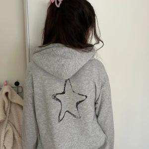 Grå hoodie från Gina Tricot med tryck - Grå hoodie från Gina Tricot med svart stjärntryck på ryggen och texten 'JE T'AIME' framtill. Mjuk insida och huva, perfekt för chill dagar. Långärmad och loose passform för en avslappnad vibe.