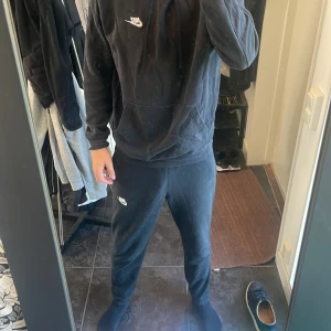 Nike tracksuit svart. - Svart hoodie från Nike med vit logga på bröstet och svarta Nike byxor med vitt Nike märke. Tröjan har huva med snörning, stor magficka och ribbade muddar vid ärmslut och nederkant. Perfekt för en chill kväll eller ett gympass, klassisk stil och mjukt material. Perfekt att få med hela setet, billigaste på marknaden. Fullt äkta!