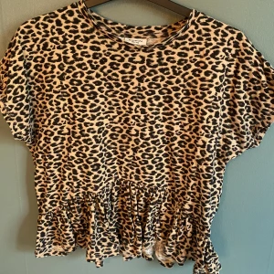 Leopardmönstrad topp med volang Kappahl - Säljer en kortärmad topp från Kappahl i storlek 146/152. Toppen har ett coolt leopardmönster i beige och svart, rund halsringning och en snygg volangdetalj längst ner. Perfekt för dig som vill sticka ut med en trendig look.
