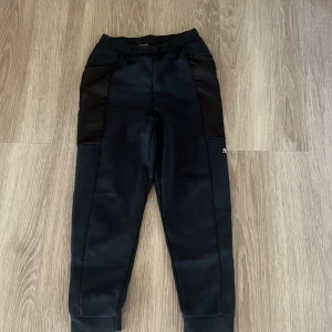 Svarta träningsbyxor från Puma - Svarta träningsbyxor från Puma med elastisk midja, dragkedjefickor och snygga paneler i mörkare svart. Byxorna har Puma-logga på benet och är gjorda i ett funktionsmaterial som andas. Perfekta för sport eller chill. Passar för 10-12 år i storlek 146-152