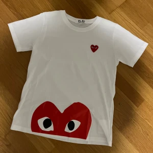 CDG Tröja - Snygg vit t-shirt från Comme des Garçons Play i bomull. Framsidan har det ikoniska röda hjärtat med ögon broderat på bröstet och ett stort tryckt hjärta med ögon längst ner. Klassisk rund hals och korta ärmar, perfekt i en clean outfit
