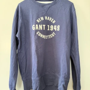 Blå sweatshirt från Gant med tryck - Säljer en blå sweatshirt från Gant med trycket 'New Haven Gant 1949 Connecticut' i vitt framtill. Tröjan har rund halsringning, ribbade muddar och är långärmad. Perfekt för en avslappnad och sportig stil.
