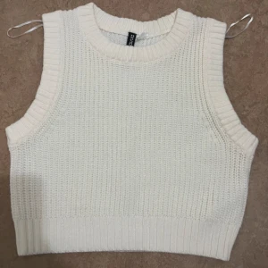 Vit stickad väst från H&M Divided - Säljer en krämvit stickad väst från H&M Divided i storlek S. Västen har breda ribbade kanter vid hals och ärmhål samt en croppad passform. Perfekt att styla över en skjorta eller t-shirt för en trendig look.