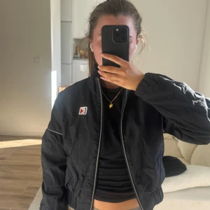 Svart vindjacka från Kani Sport XS - Svart vindjacka från Karl Kani i storlek XS. Jackan har dragkedja framtill, broderad logga på bröstet och stor KANI SPORT-text på ryggen. Lätt material med meshfoder och reflekterande detaljer längs dragkedjan och ärmarna. Perfekt för en sportig och chill stil.