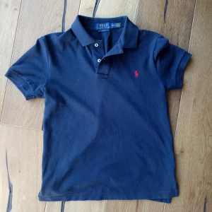 Marinblå pikétröja från Ralph Lauren - Klassisk marinblå pikétröja från Ralph Lauren med korta ärmar och krage. Tröjan har två knappar vid halsen och den ikoniska röda polospelaren broderad på bröstet. Perfekt för en stilren och sportig look.Den är äkta och är köpt på NK.