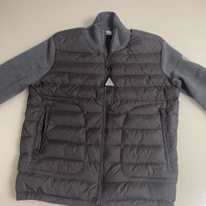 Grå dunväst från Moncler med stickade ärmar - Snygg grå dunväst från Moncler med stickade grå ärmar och hög krage. Västen har dragkedja framtill och två dragkedjefickor. Klassisk Moncler-logga på ärmen. Perfekt för lager-på-lager och stilren look.