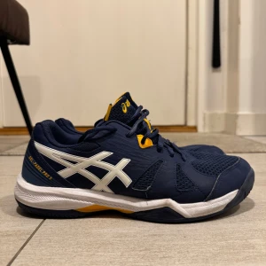 Blå Asics Gel-Padel Pro 5 sneakers - Snygga Asics Gel-Padel Pro 5 sneakers i marinblått med vita och gula detaljer. Ovandelen är i mesh och syntet för bra andningsförmåga och komfort. Skorna har snörning och en stabil, platt sula som passar perfekt för padel eller annan träning. Helt nya!