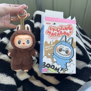 Brunt Toffee nyckelringshänge Pop Mart - Söt Toffee-figur från Pop Mart Exciting Macaron-serien, klädd i en fluffig brun dräkt med kaninöron och ett brett leende. Nyckelringen har guldigt fäste och är tillverkad av polyester och PVC. Perfekt att hänga på väskan eller nycklarna för en lekfull touch. Äkta secret labubu macaron 