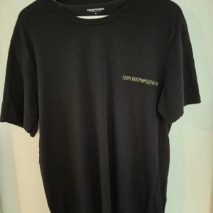 Svart t-shirt från Emporio Armani XL - Svart t-shirt från Emporio Armani med diskret logga på bröstet. Klassisk rund halsringning och korta ärmar. Tillverkad i mjuk bomull som känns skön mot huden. Perfekt för en clean och stilren look.