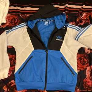 Adidas blå och svart vindjacka M - Snygg Adidas vindjacka i blått, svart och vitt med klassiska tre ränder på ärmarna. Jackan har huva, dragkedja framtill och två fickor med dragkedja. Materialet är lätt och andas, perfekt för blåsiga dagar. Adidas-logga broderad på bröstet.