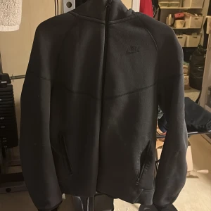 Svart Nike hoodie med zip - Säljer en svart hoodie från Nike i storlek 170. Tröjan har hel dragkedja framtill, två fickor med dragkedja och en klassisk huva. Perfekt för chill dagar eller när du vill ha en sportig look. Materialet är mjukt och bekvämt.