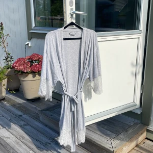 Ljusgrå morgonrock med spetsdetaljer - Superfin ljusgrå morgonrock med knytband i midjan och vacker spetsdetaljer vid ärmslut och nederkant. Morgonrocken har trekvartsärmar och är tillverkad i mjukt, tunt material som känns skönt mot huden. Perfekt för en chill och avslappnad look.