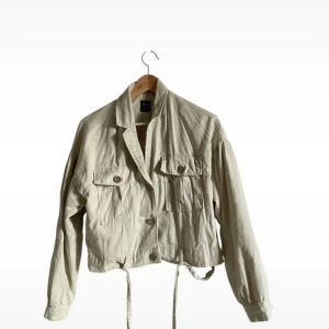 Beige jacka - Säljer en beige jacka med knappar framtill, två stora fickor och knytband i midjan. Jackan har lång ärm och en avslappnad passform, perfekt att slänga på över en t-shirt. Materialet är bomull och den har en stilren look.