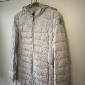 Ljusbeige dunjacka Parajumpers S - Lätt och snygg ljusbeige dunjacka från Parajumpers med huva och dragkedja framtill. Jackan har quiltad design, innerficka med 'security pocket'-detalj och klassisk PJS-logga på ärmen. Perfekt för kyliga dagar när du vill hålla stilen.