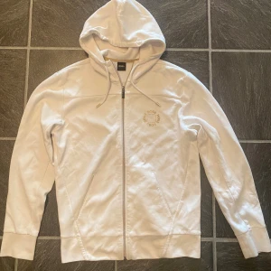Vit hoodie från BOSS med dragkedja - Vit hoodie från BOSS med dragkedja framtill och snörning i huvan. Tröjan har fickor på sidorna och en diskret BOSS-logga i guld på bröstet. Finns med matchande underdel (dress)