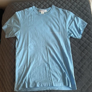 Ljusblå t-shirt från Comme des Garçons - Säljer en stilren ljusblå t-shirt från Comme des Garçons SHIRT. T-shirten är i 100% bomull, har rund halsringning och diskret tryck på bröstet. Enkel och clean design som passar perfekt till jeans eller shorts.