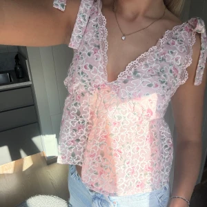 Rosa blommig spetsblus med knyt - Supersöt blus i ljusrosa spets med blommigt mönster och vita broderade detaljer. V-ringad med spetskant och knytband på axlarna. Luftig och lätt, perfekt till jeans eller kjol. Blusen har en loose passform och är ärmlös.