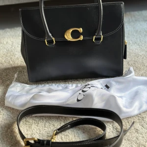 Svart handväska från Coach i skinn - Säljer en stilren svart handväska från Coach i slätt skinn med guldiga metalldetaljer och stor C-logga framtill. Väskan har både handtag och avtagbar axelrem, samt brunt innerfoder. Perfekt för dig som vill ha en klassisk och snygg accessoar.