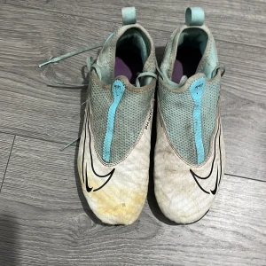 Nike Phantom GT2 fotbollsskor ljusblå - Nike Phantom GT2 fotbollsskor i ljusblått och vitt med svarta detaljer. Skorna har snörning, meshpaneler för andning och dobbar för bra grepp på planen. Swoosh-logga på sidan och dragflik bak. Perfekta för dig som vill sticka ut på fotbollsplanen.